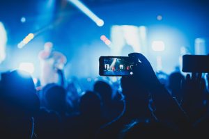 Photo of a crowded concert with a phone live streaming the event in the foreground. The lights are mostly blue.
